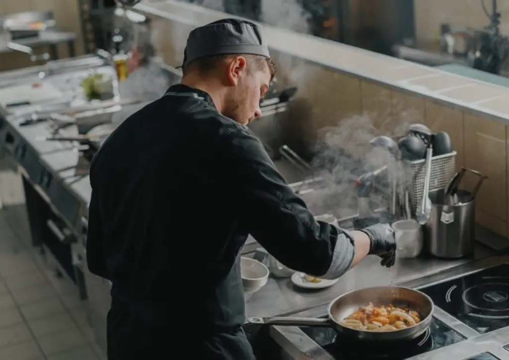Professional chef in black uniform and gloves seasoning food in a frying pan on a commercial induction stove in a restaurant kitchen