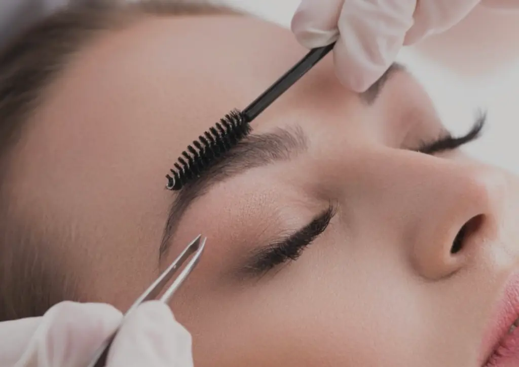 Beautician in white gloves shaping and grooming a client's eyebrow using a spoolie brush and tweezers during a salon treatment