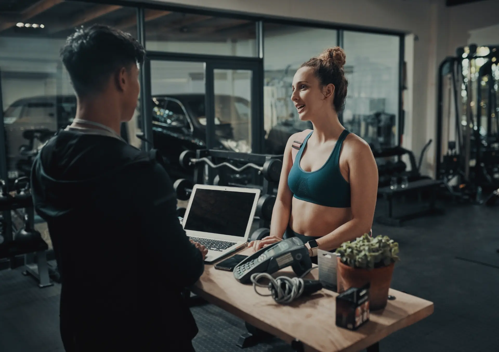 A gym staff member assisting a client at the reception desk with a laptop and payment terminal