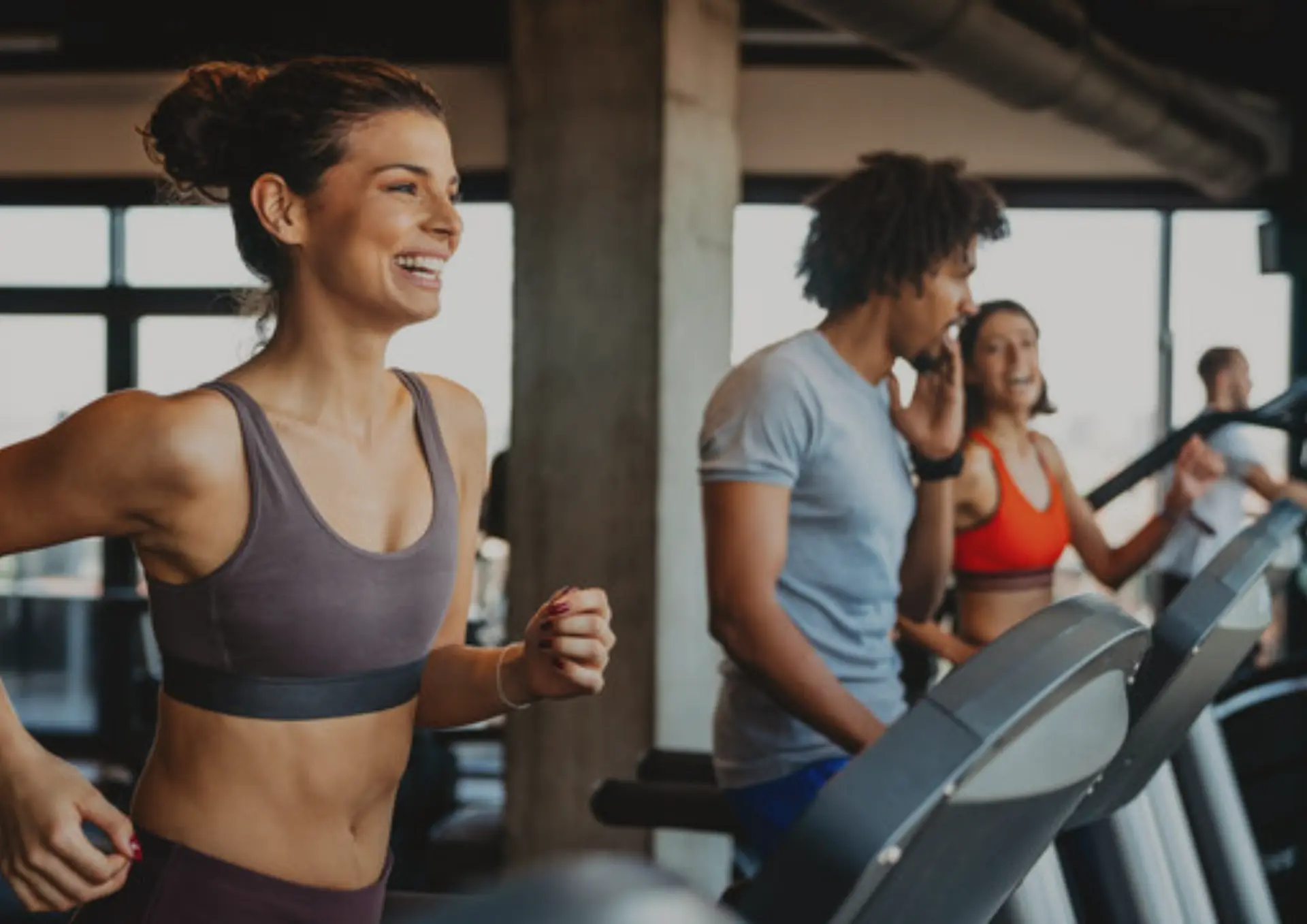 A smiling woman running on a treadmill in a busy gym with other people exercising in the background
