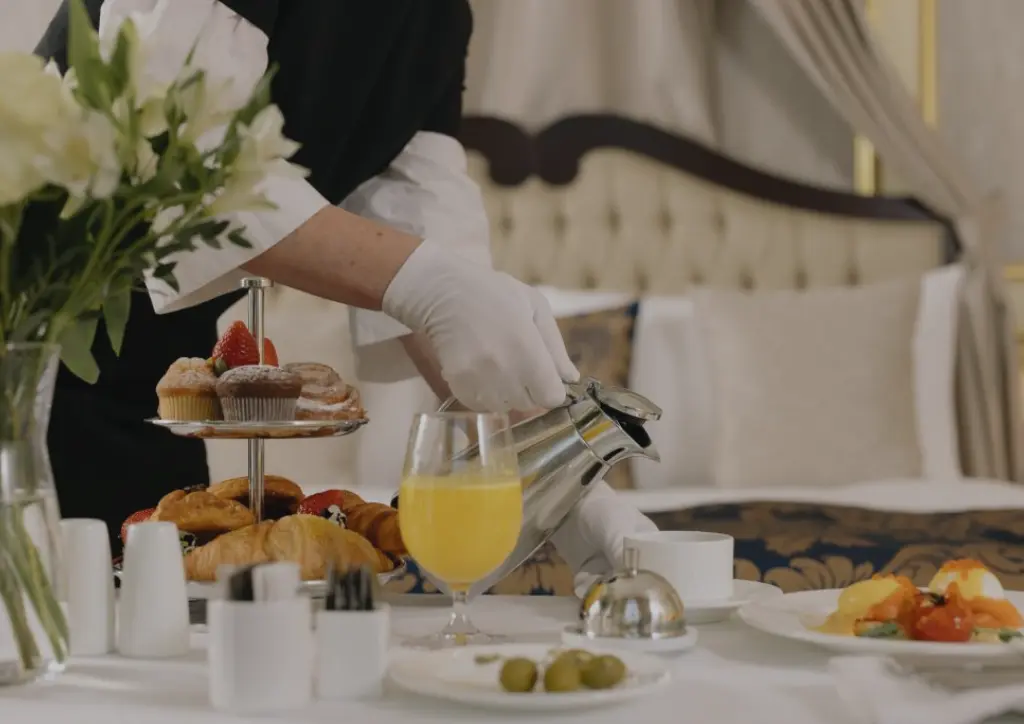 Hotel room service staff in white gloves pouring a hot drink for a luxury breakfast spread with pastries, orange juice and eggs benedict