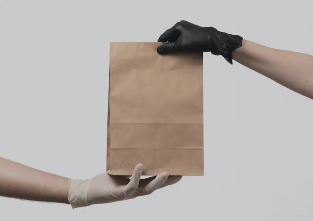 A person wearing a black glove handing over a brown paper takeaway food delivery bag to a person wearing a white glove against a plain white background